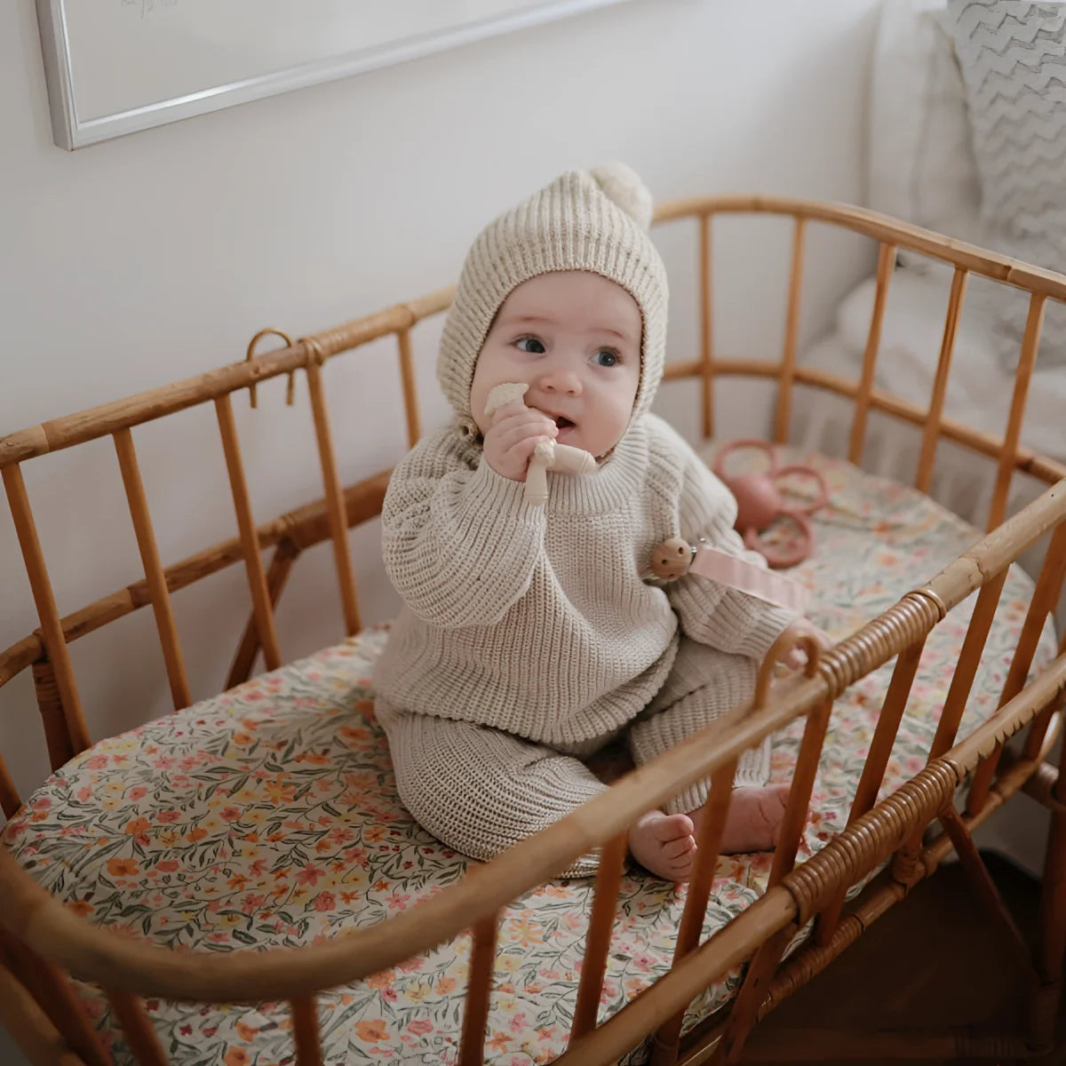 Baby im Holzbett in gestricktem Creme-Outfit und Mütze neben Silikon Lamm Beisstier