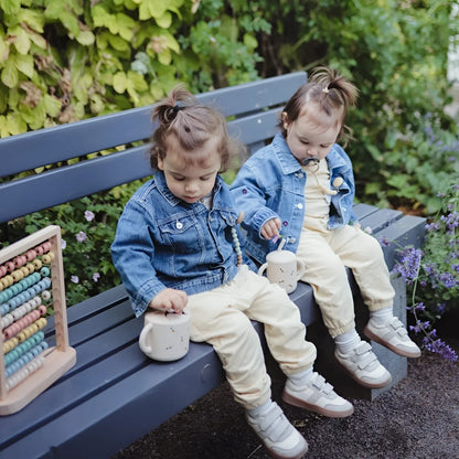 Zwei kleinkinder in blauen jeansjacken und cremefarbenen hosen sitzen mit weißen keramik snack bechern bee auf einer bank