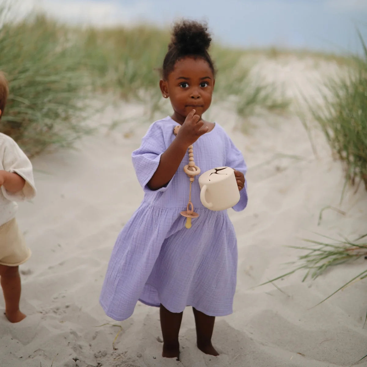 Junges Kind im lila Kleid steht auf Sand neben Snack Becher Shifting Sand