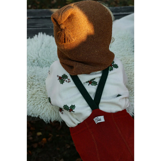 Braune Strickmütze mit Bommel, weißer Pullover mit Stechpalmenprint, rote Hosenträger