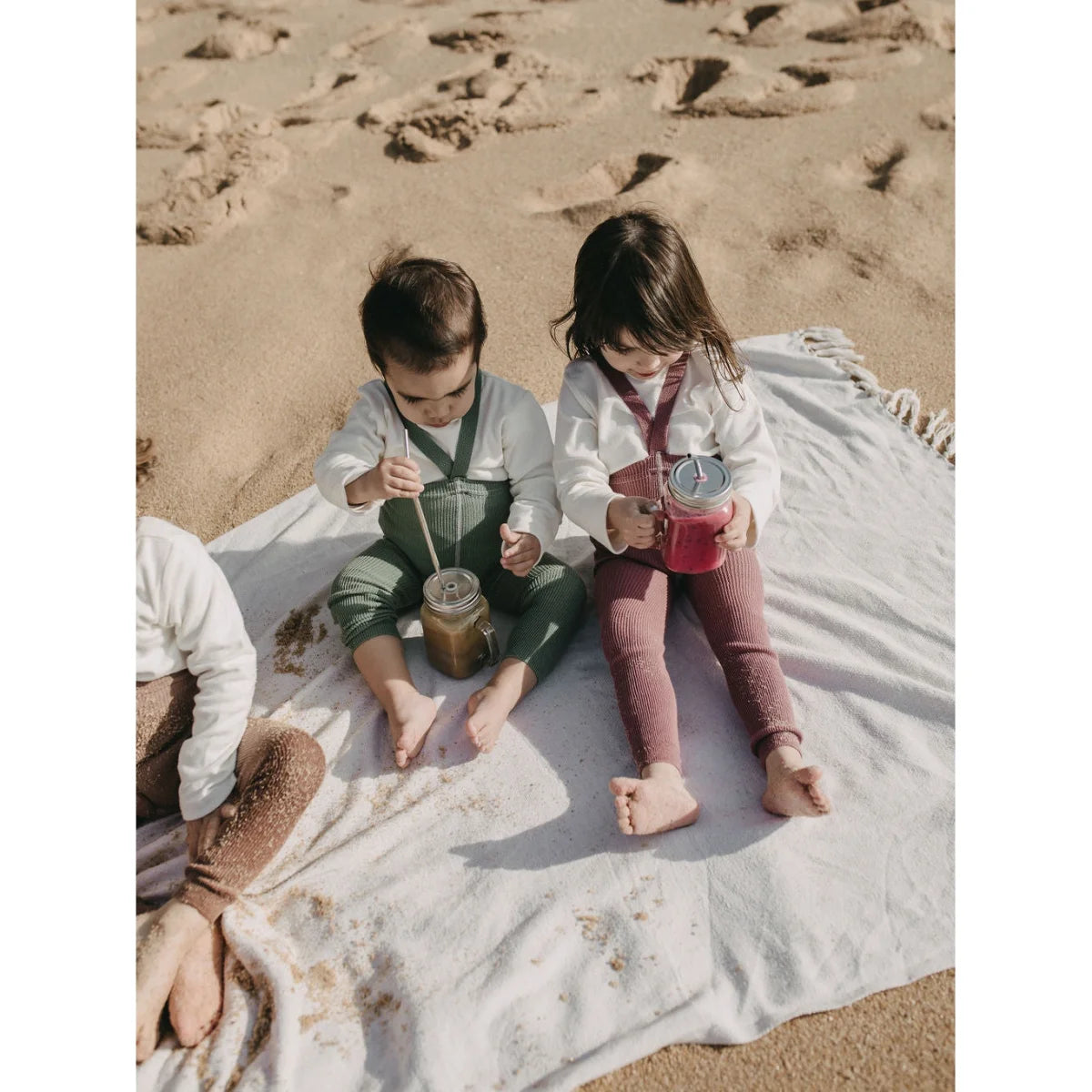 Zwei Kinder sitzen am Strand auf einer Decke und halten Getränke