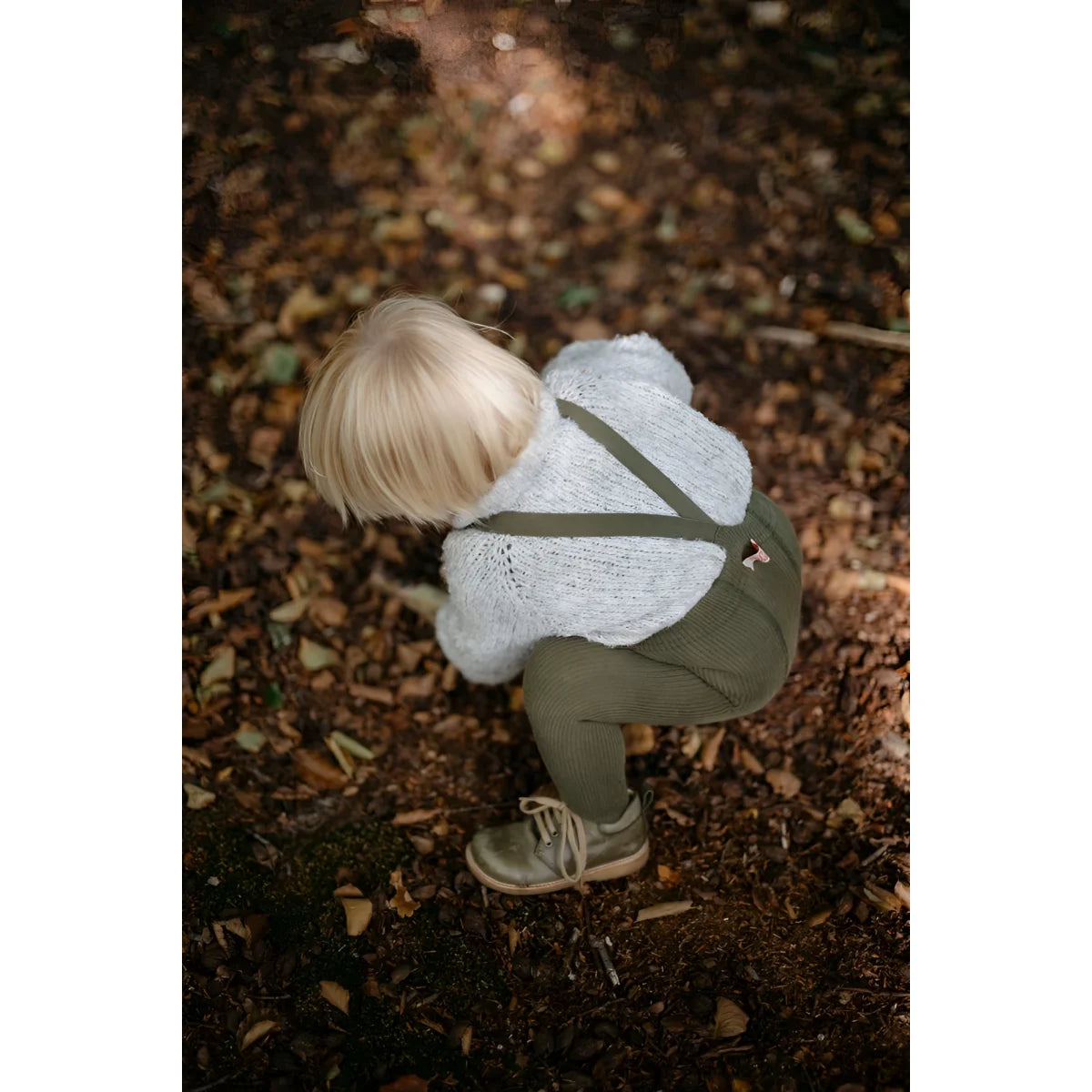 Kind hockt auf dem Boden in weißem Pullover und grüner Latzhose, Strumpfhose mit Träger