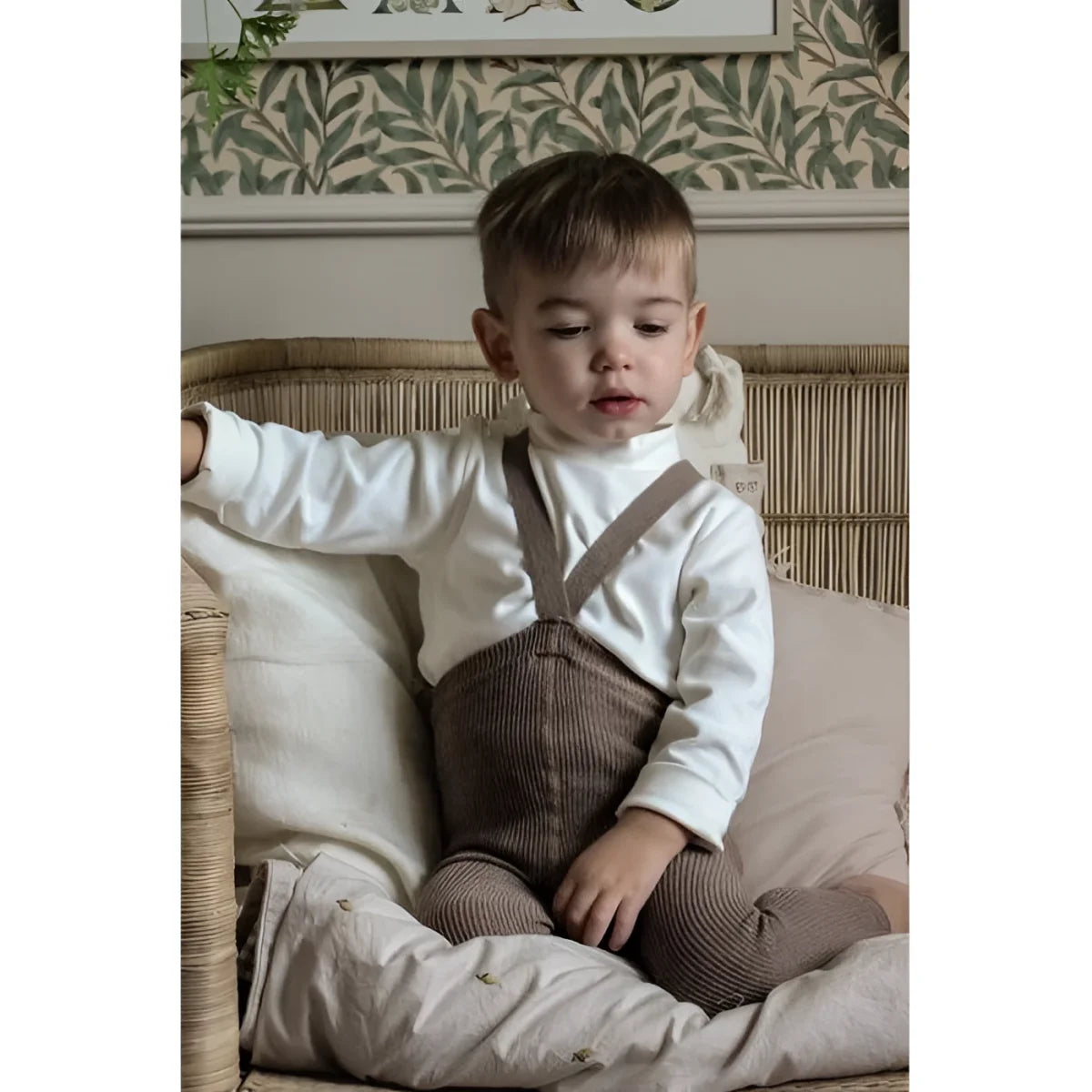 Junger Kind in weißem Pullover und braunen Strickoveralls auf Kissen sitzend