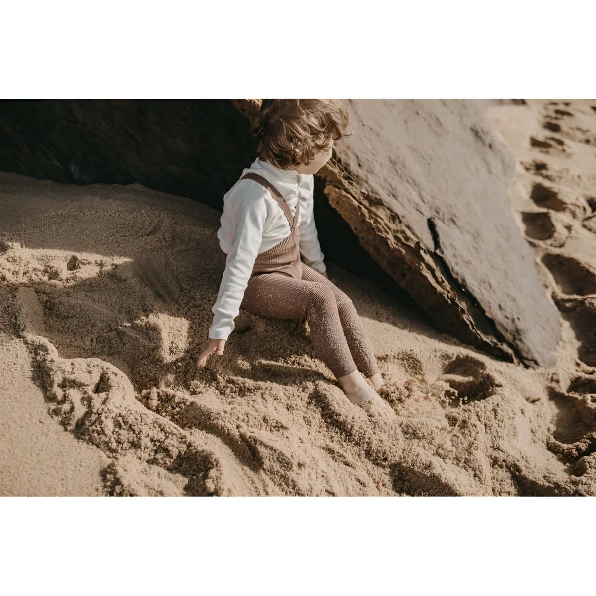 Kind sitzt am Sandstrand neben Felsen in Strumpfhose mit Träger Footless | Granola