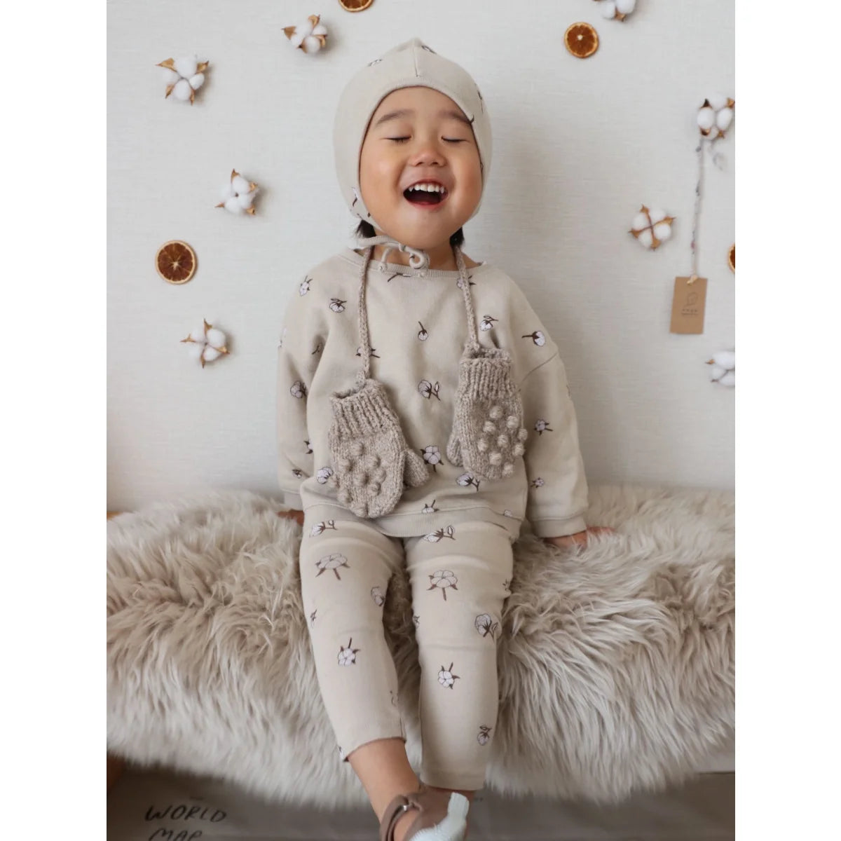 Lachendes Kind in cremefarbigem Outfit mit Mütze und Handschuhen für Cottonfield Pullover