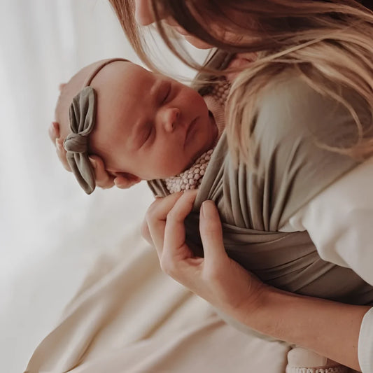 Neugeborenes Baby mit Schleifenhaarband in den Armen einer Person, Tragetuch Dusty Olive