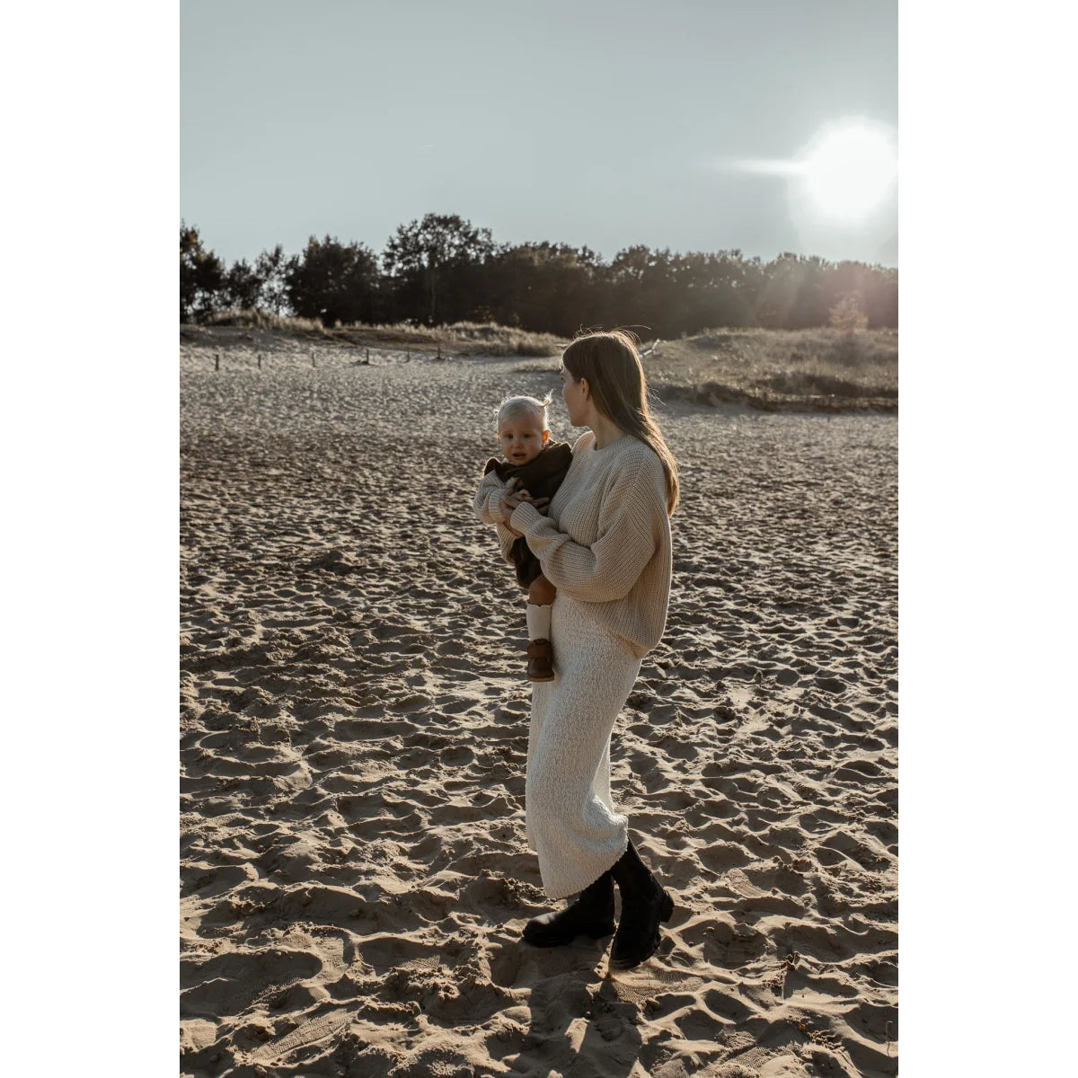Frau hält Baby am Strand bei Sonnenuntergang in einem grob gestrickten Pullover