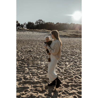 Frau hält Baby am Strand bei Sonnenuntergang in einem grob gestrickten Pullover