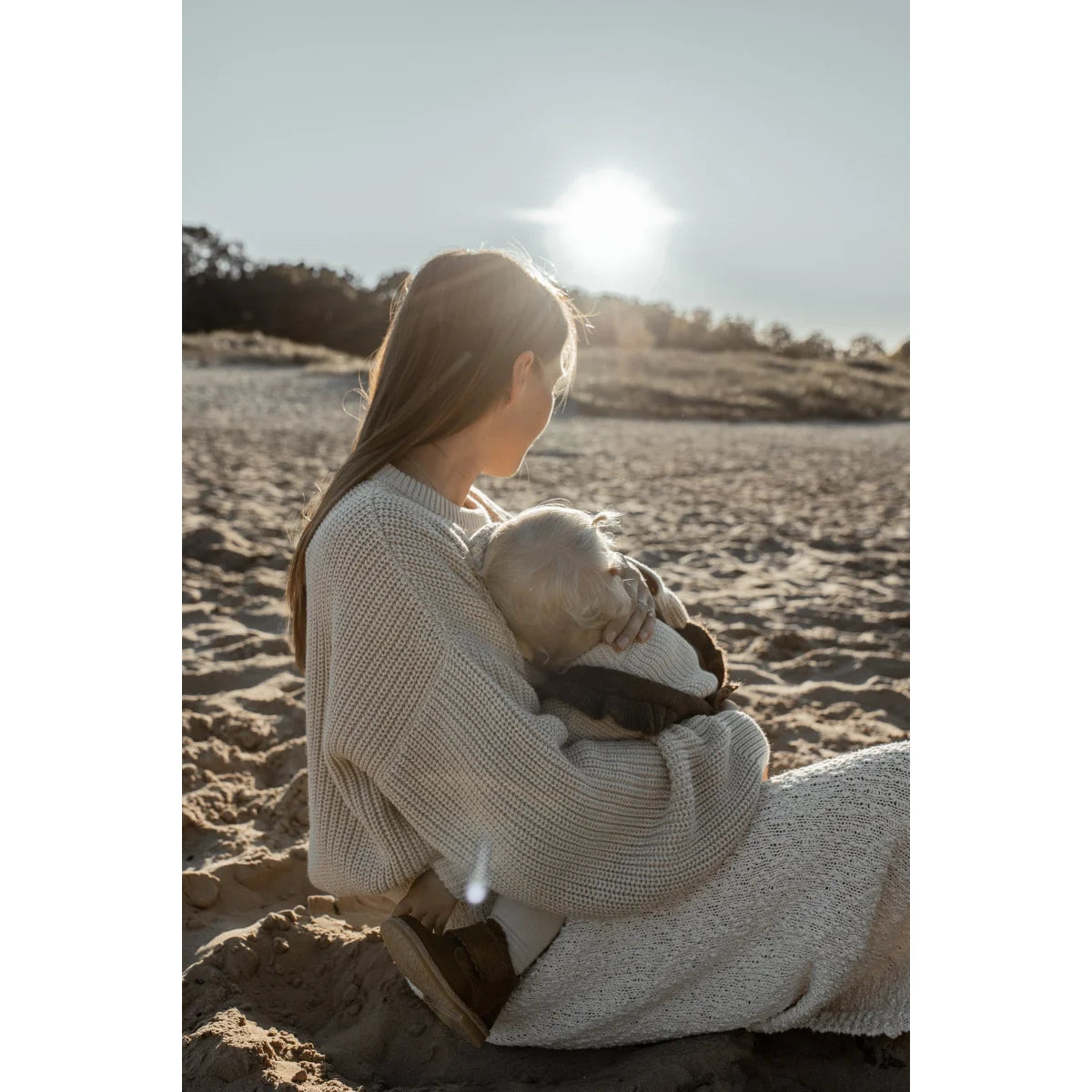Frau mit Baby am Strand bei Sonnenuntergang in einem grob gestrickten Pullover