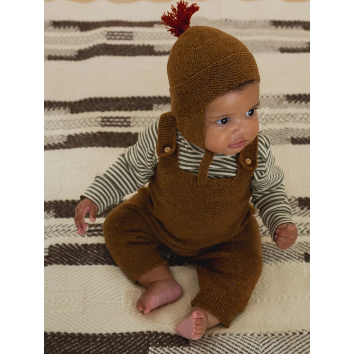 Niedliches Baby trägt braune Strickmütze mit rotem Zopf und passende Overalls